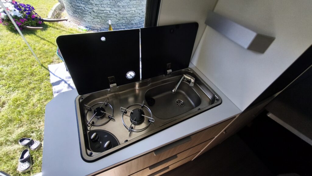 Obrázek zobrazuje kuchyňský kout v obytném autě s dřezem a dvouplotýnkovým vařičem.