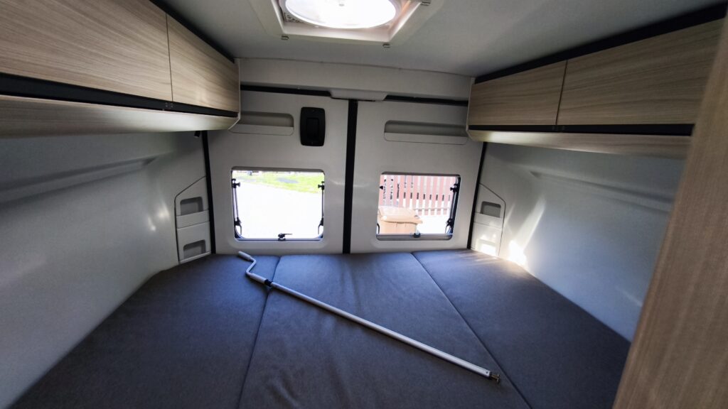 Obrázek interiéru karavanu, světlé dřevěné skříňky a prostorná postel pro pohodlné spaní.
