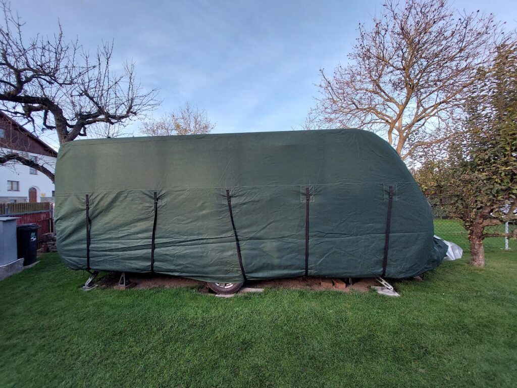 Na obrázku je zakrytý karavan, uložený na trávě. Není vidět značka ani model.