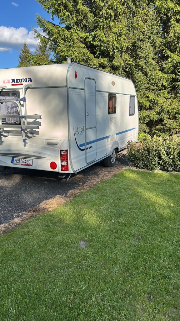 Karavan Adria Altea 432 PH parkovaný na zelené trávě, obklopený stromy, ideální pro kempování.