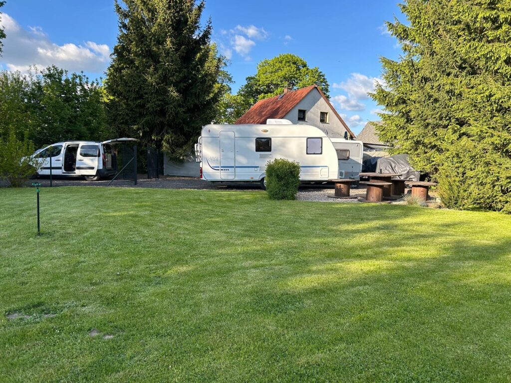 Na obrázku je karavan zaparkovaný na zahradě, vedle obytňáku. Vzadu je stříšková konstrukce a stůl s lavičkami.