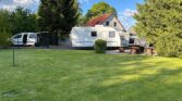 Na obrázku je karavan zaparkovaný na zahradě, vedle obytňáku. Vzadu je stříšková konstrukce a stůl s lavičkami.