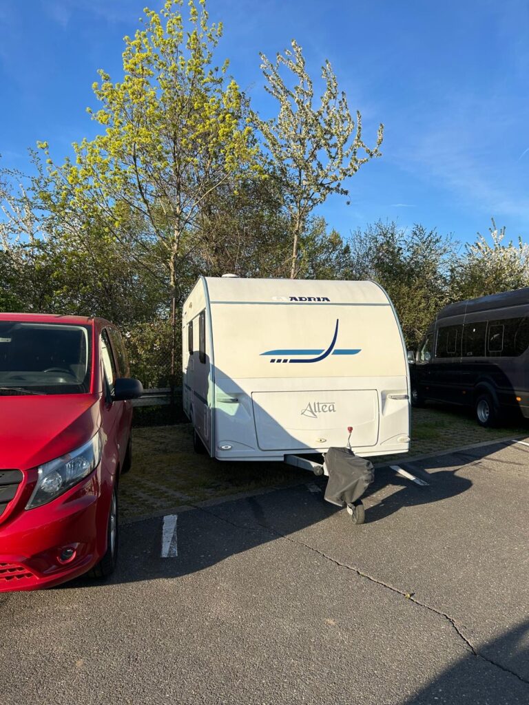 Karavan Adria Altea zaparkovaný mezi stromy, vedle červeného auta. Ideální pro pohodlné cestování!