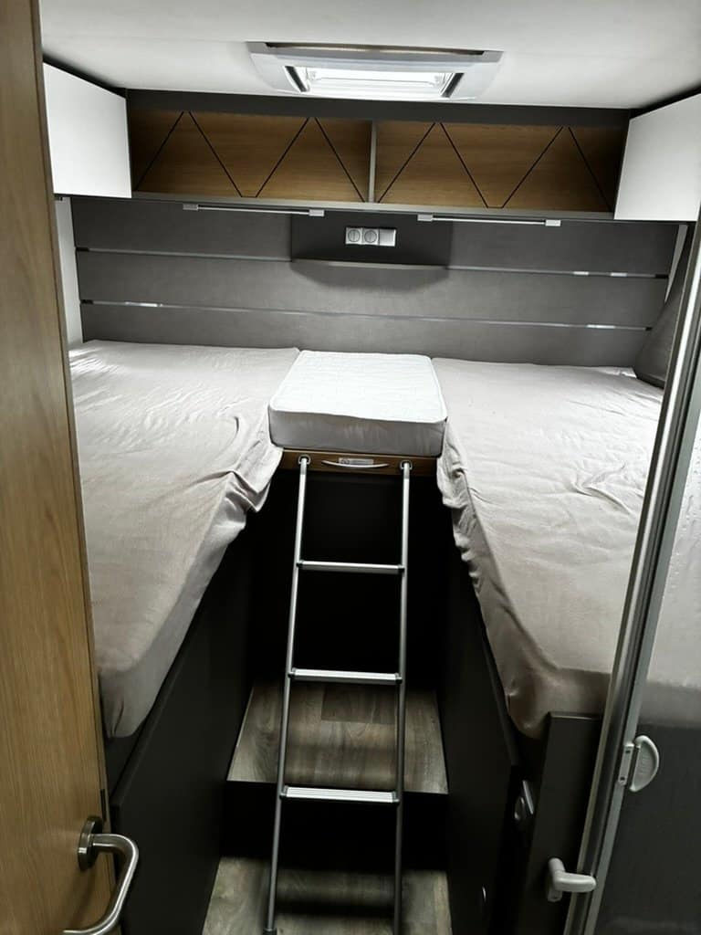Interiér obytného auta s dvoulůžkem, schůdky a úložným prostorem. Moderní design a pohodlí na cestách.