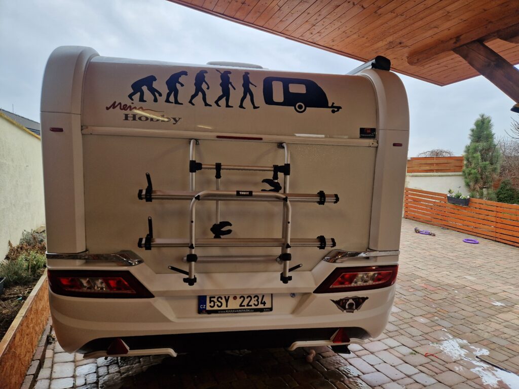 Na zadní straně bílého obytného vozu je nálepka zobrazující vývoj člověka vedoucí k někomu, kdo táhne karavan, což naznačuje prodej karavanu. Dodávka je zaparkovaná pod dřevěným přístřeškem na dlážděné příjezdové cestě.
