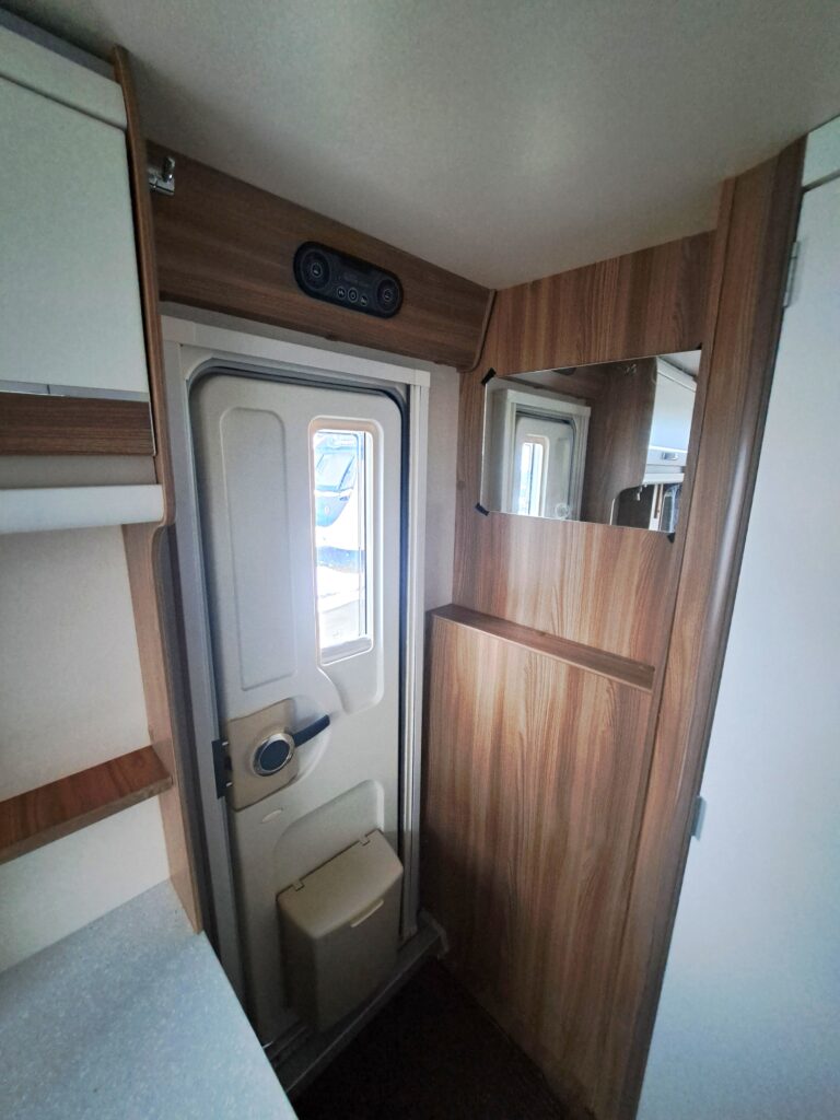 Obrázek interiéru karavanu s dřevěným obložením a vstupními dveřmi. Značka a model nejsou uvedeny.