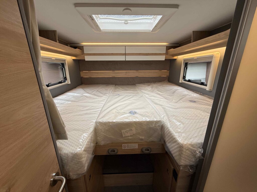 Obrázek interiéru obytného auta s dvojlůžkem, moderní design a prostorné úložné prostory. Ideální pro pohodlné cestování.