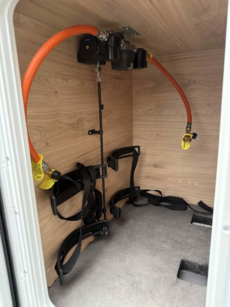 Obrázek ukazuje prostor pro připojení v obytném autě, s potrubím a úchyty pro vybavení.