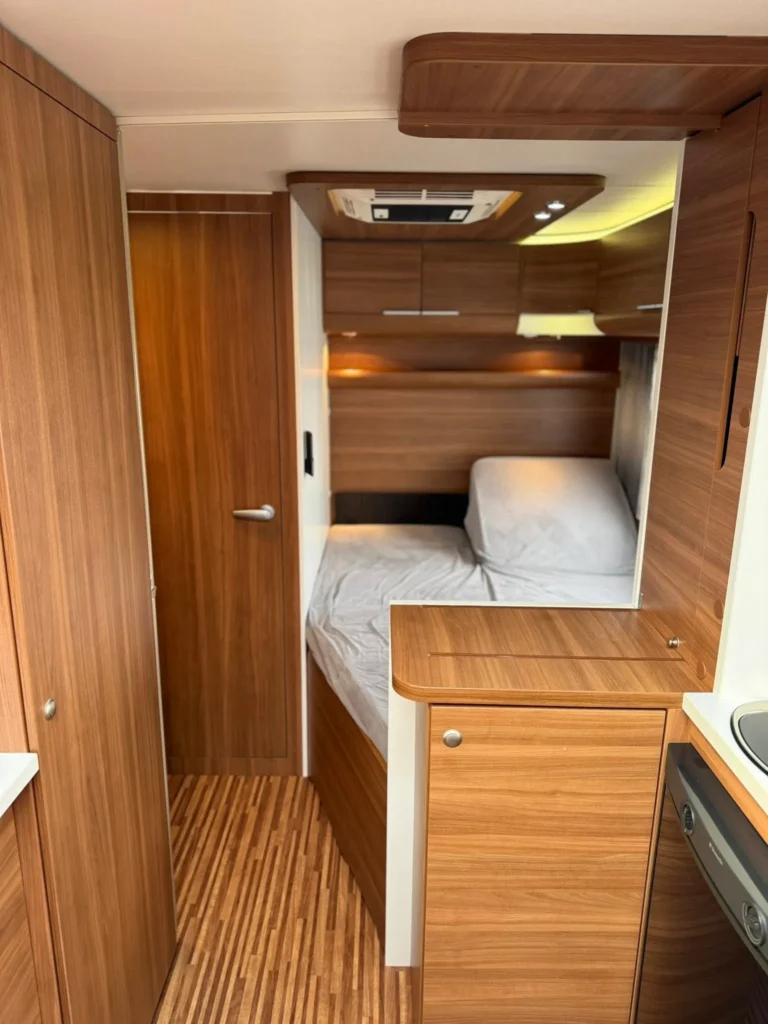 Obrázek interiéru moderního karavanu s dřevěnými prvky. Prostorné uspořádání s dvojlůžkem a kuchyňským koutem.