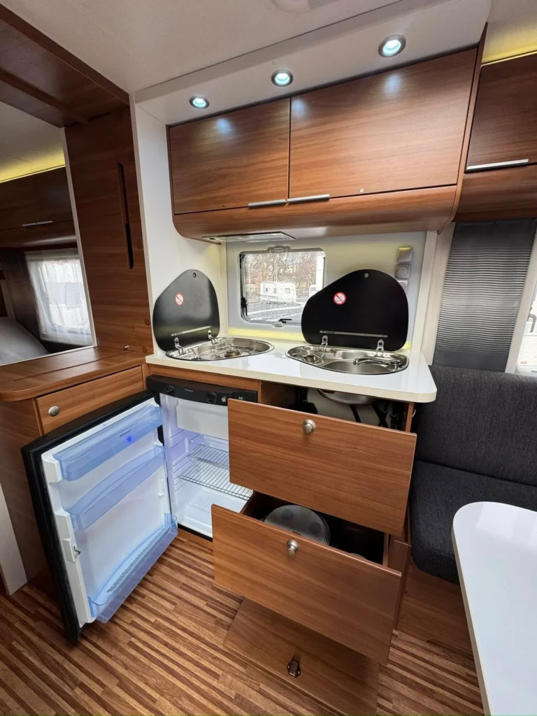 Obrázek interiéru karavanu s moderní kuchyní, lednicí a úložnými prostory. Komfortní a praktický design pro cestování.