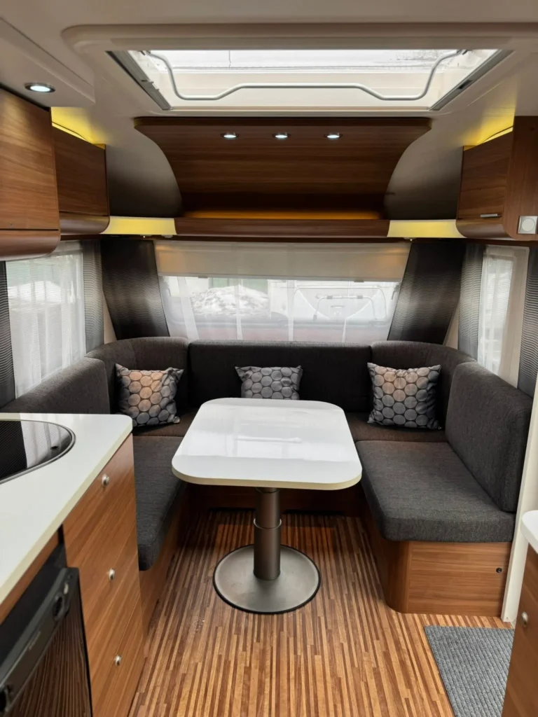 Interiér moderního obytného přívěsu s kuchyňským koutem, útulným sezením a stolečkem. Decentní dřevěné prvky.