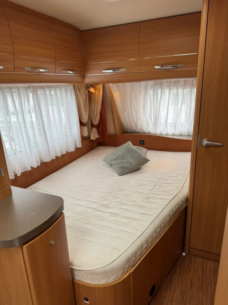 Obrázek interiéru karavanu s prostornou postelí a úložným prostorem. Příjemné dřevo a světlo vytváří útulnou atmosféru.