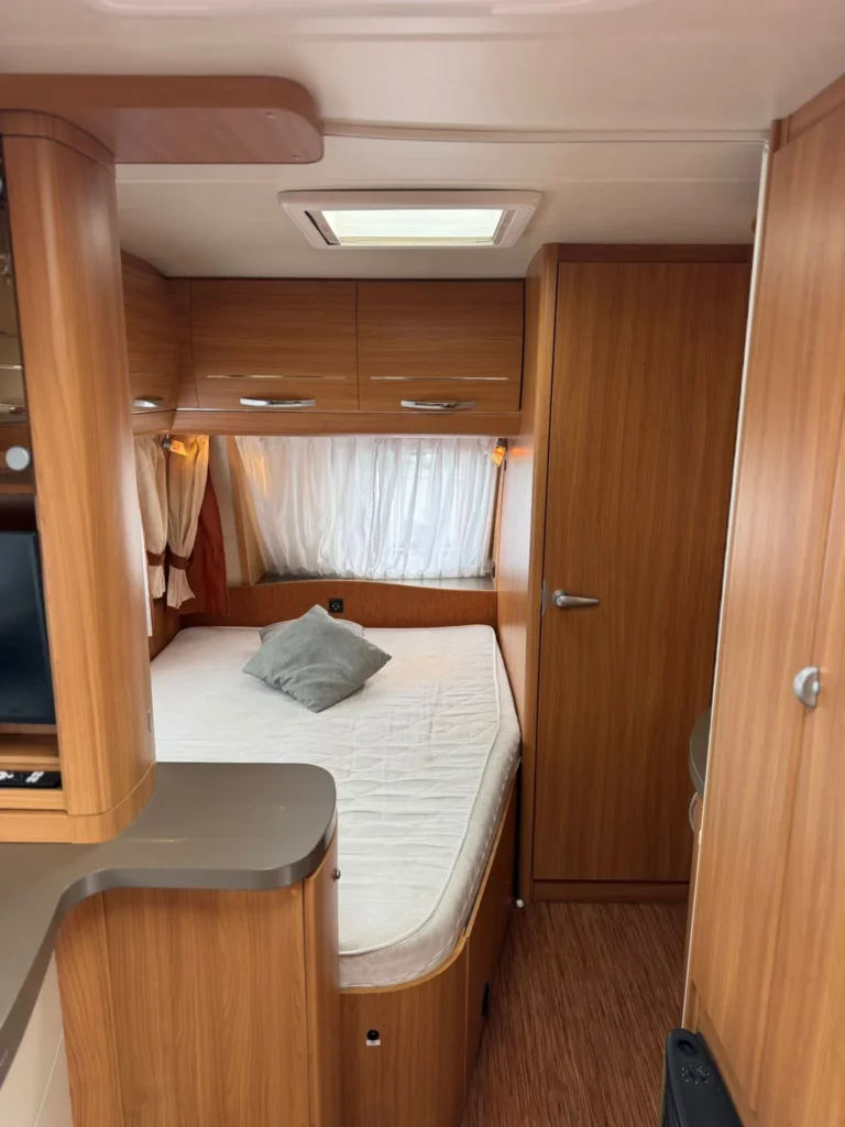 Interiér karavanu s dřevěnými prvky, pohodlnou postelí a úložnými prostory. Idealní pro kempování a cestování.