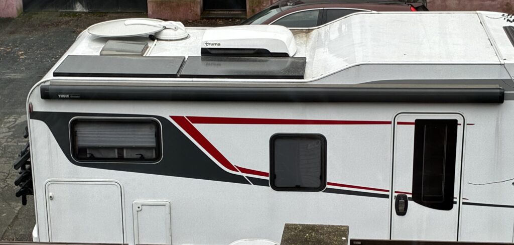 Na obrázku je karavan s bílým exteriérem, červenými a černými pruhy. Viditelný je střešní ventilátor a markýza Thule.