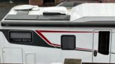 Na obrázku je karavan s bílým exteriérem, červenými a černými pruhy. Viditelný je střešní ventilátor a markýza Thule.