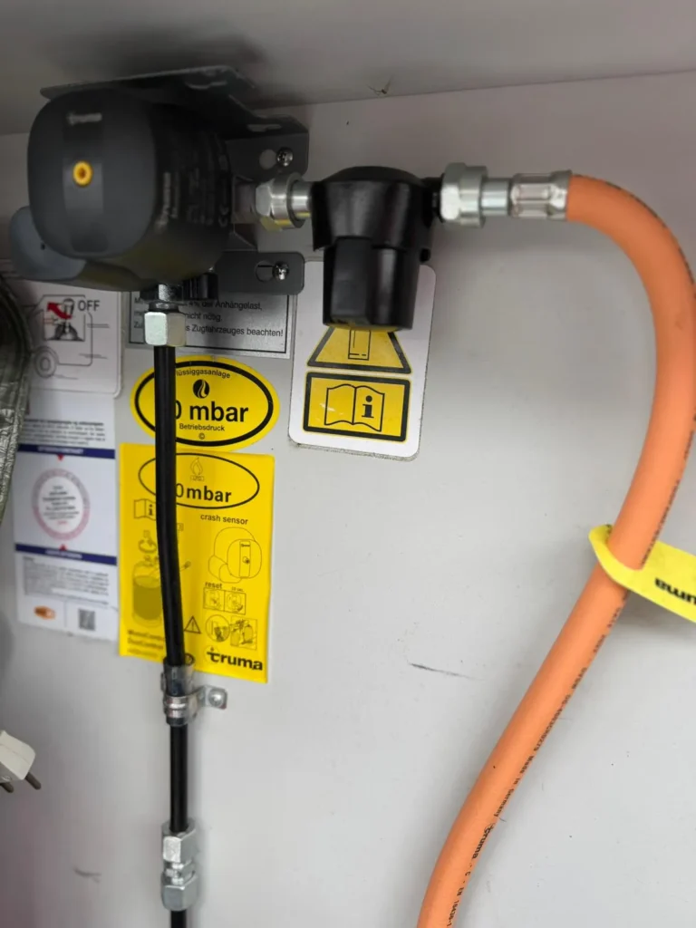 Obrázek ukazuje zařízení Truma s hadicemi a bezpečnostními značkami, které se používají v obytných autech a karavanech.