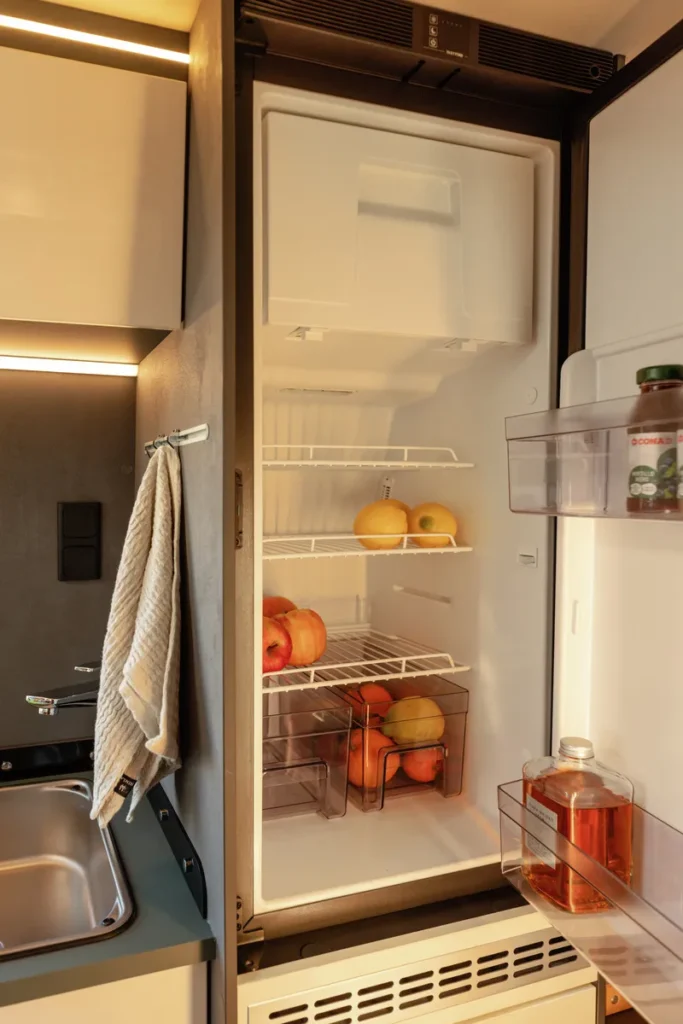 Obrázek zobrazuje interiér obytného vozu s chladničkou plnou ovoce a nápojů.