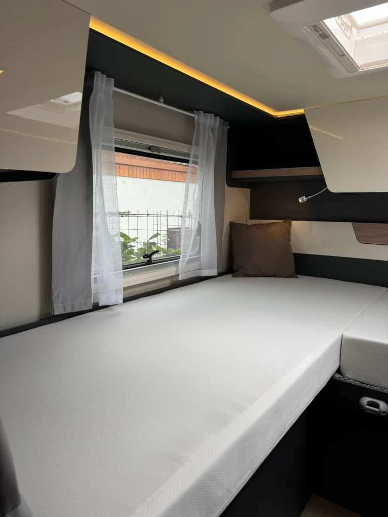 Interiér karavanu s bílým lůžkem, světlými závěsy a moderním designem. Vidět část okna a osvětlení.