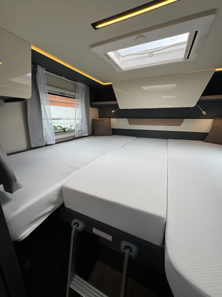 Interiér obytného přívěsu s bílými matracemi a moderním osvětlením. Vidět okno a úložné prostory.