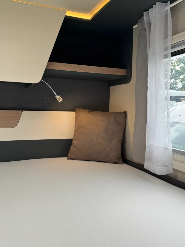 Interiér karavanu s bílou sedačkou a hnědým polštářem, viditelné úložné prostory a okno s bílou záclonou.