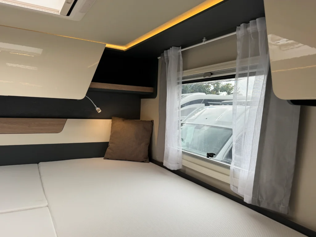 Interiér moderního karavanu s manželskou postelí, úložnými skříňkami a okny s bílými záclonami.