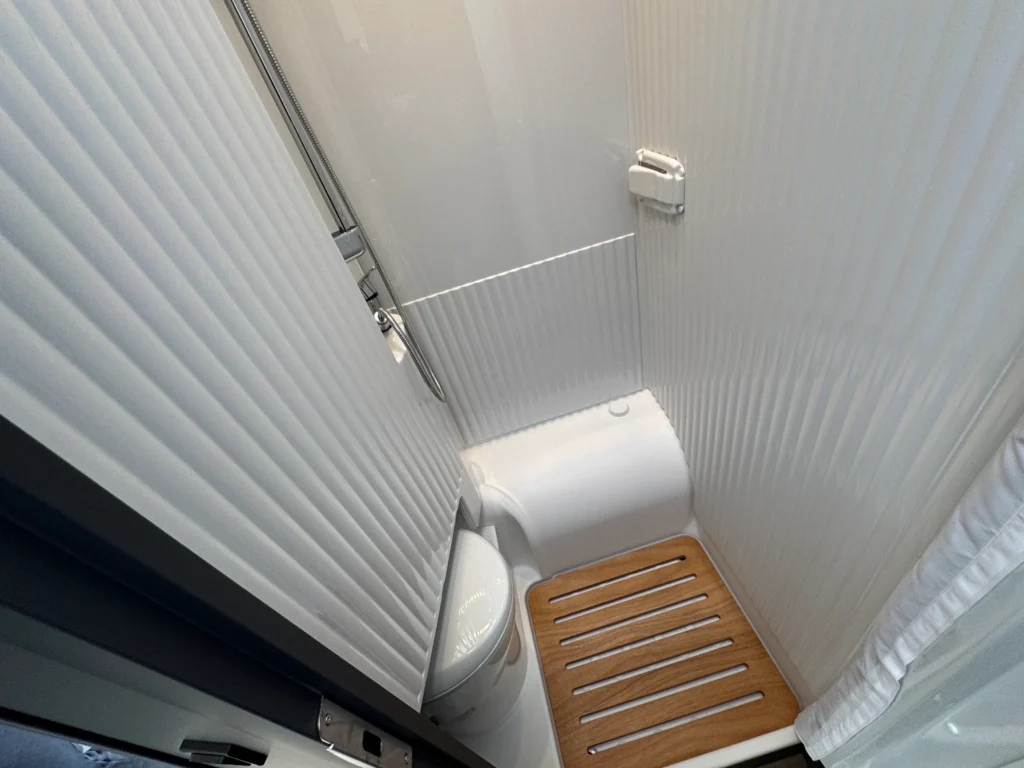 Obrázek ukazuje moderní koupelnu obytného auta s toaletu a sprchou, minimalistické bílé provedení.
