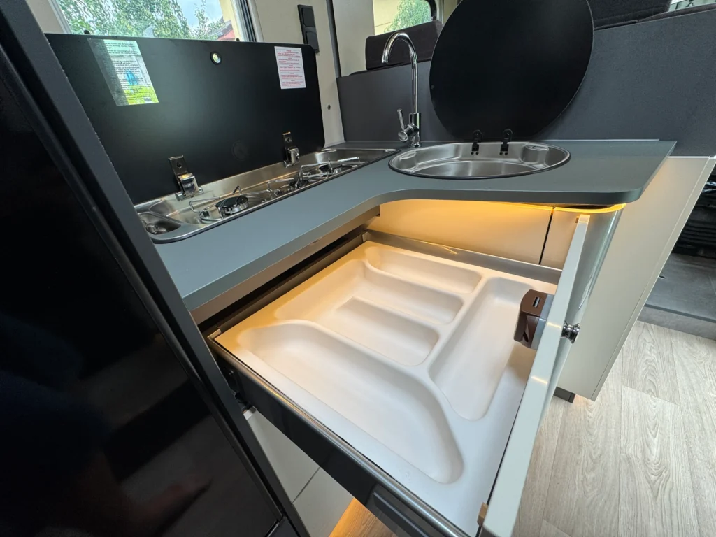 Obrázek ukazuje moderní kuchyňku v obytném autě s dřezem a dvěma vařiči. Ideální pro pohodlné vaření na cestách.