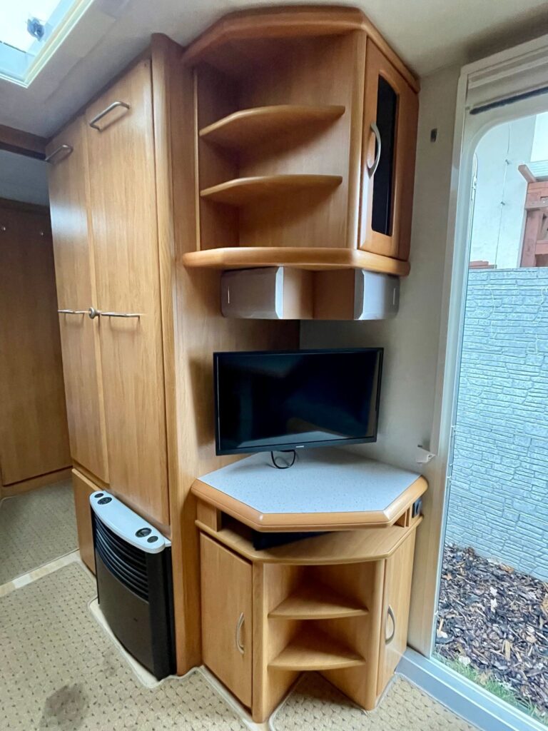 Interiér obytného auta s televizí a úložným prostorem, dřevěné prvky a komfortní design.