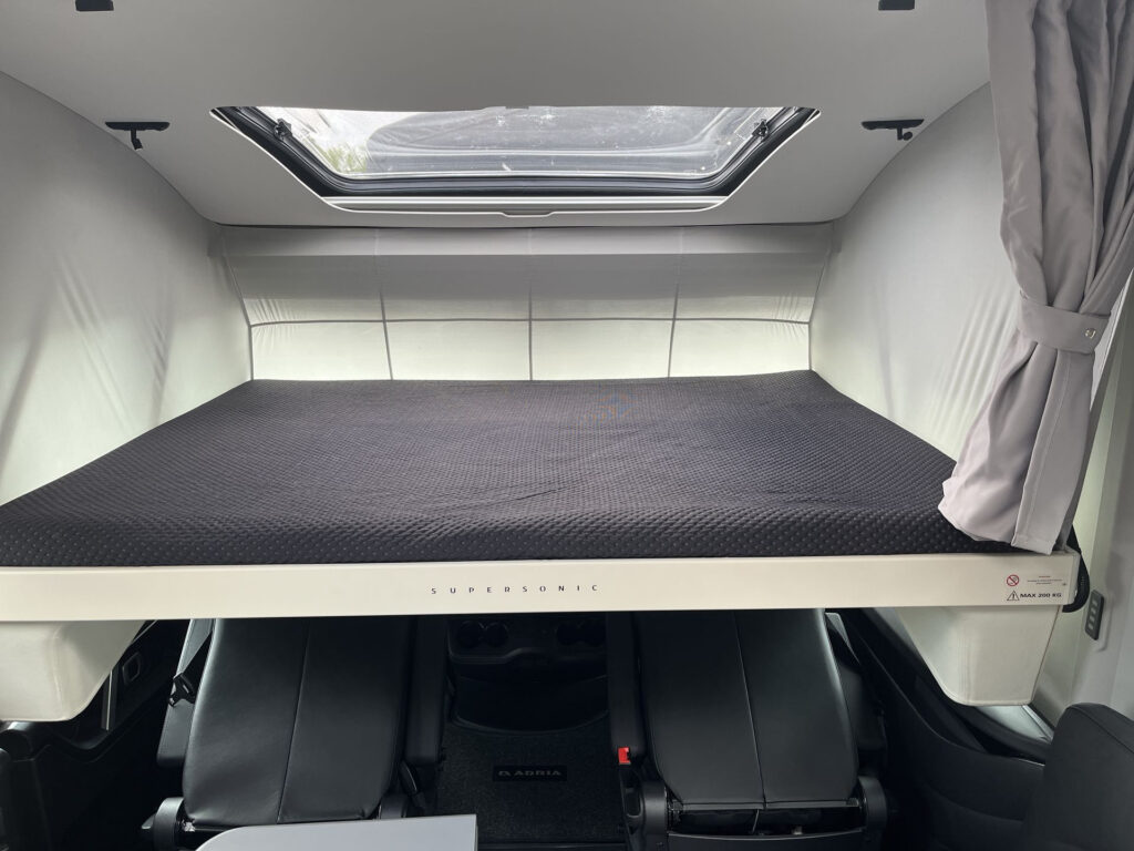 Obrázek interiéru obytného auta, výklopné lůžko a pohodlné sedadla. Vůz má značku Adria Supersonic.