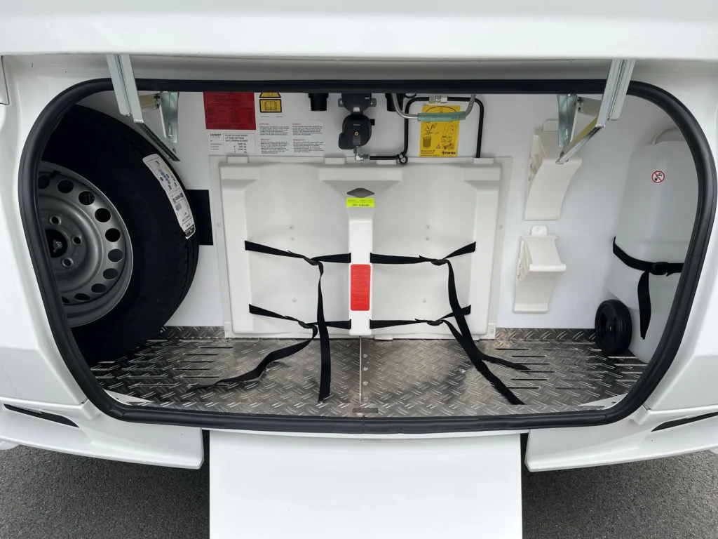 Obrázek ukazuje úložný prostor obytného auta s rezervním kolem a dalšími prvky. Značka a model nejsou vidět.