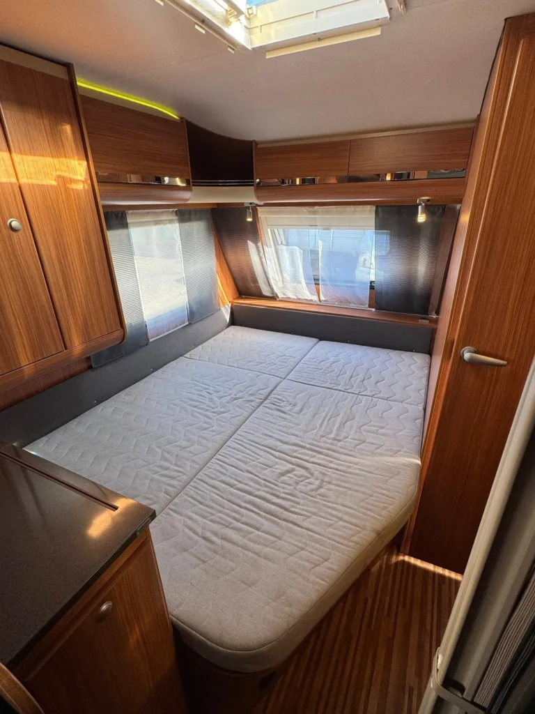 Interiér karavanu s pohodlným dvojlůžkem a moderním dřevěným nábytkem. Ideální pro pohodlný odpočinek při cestování.