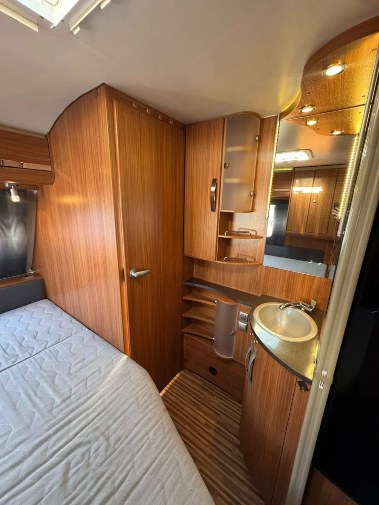 Interiér přívěsu s dřevěnými prvky, pohodlnou postelí a koupelnou. Ideální pro příjemné cestování.