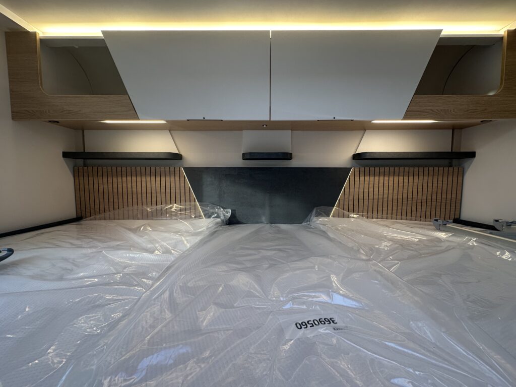 Obrázek interiéru s lůžkem a úložnými prostory v obytném automobilu, bez viditelné značky nebo modelu.