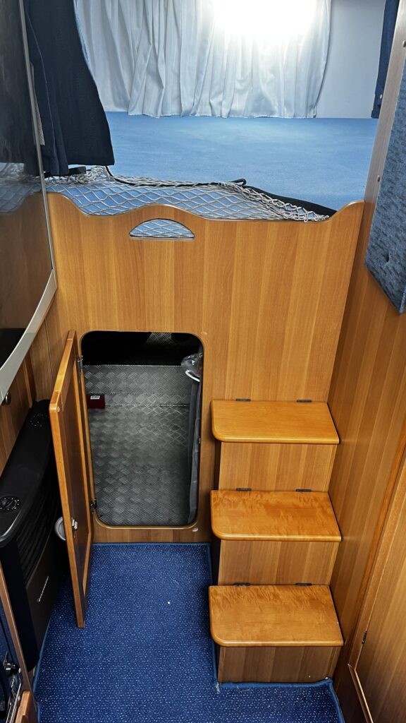 Interiér obytného vozu s dřevěnými schody a úložným prostorem. Moderní a praktický design pro pohodlný pobyt.