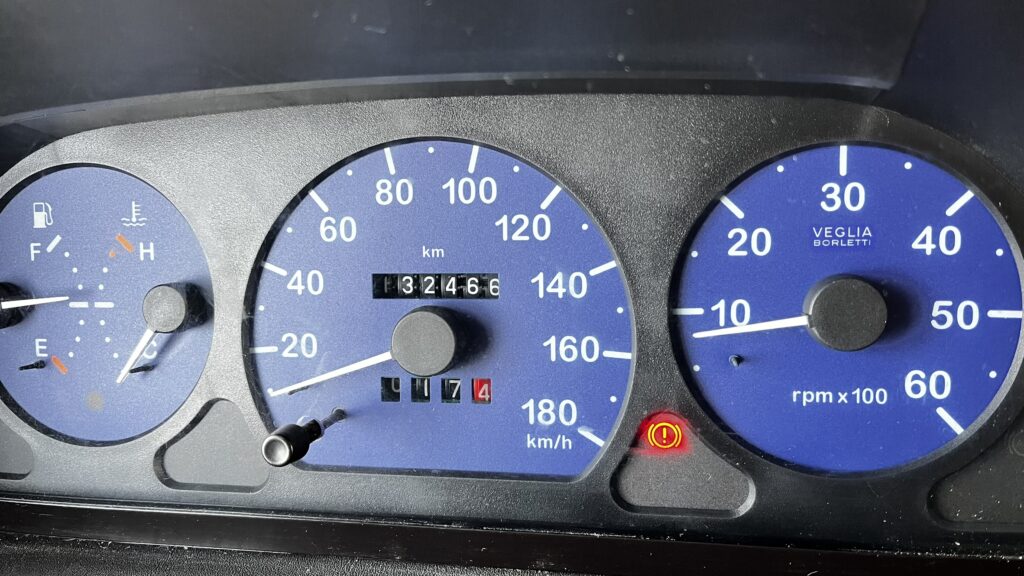 Obrázek přístrojové desky obytného auta, ukazuje tachometr a ukazatele teploty, paliva a otáček motoru.