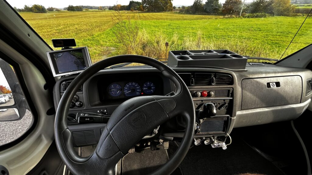 Interiér obytného auta s přehlednou palubní deskou a výhledem na zelené pole.