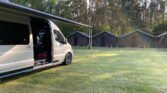Obytné auto s vytaženou markýzou v přírodě, ideální pro dobrodružství a odpočinek. V pozadí chatky.